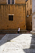 Toskana, Italien - 23. April 2025: Blick auf eine einsame Gestalt, die die sonnenüberflutete Piazza überquert, wo der warme, honigfarbene Stein der alten Gebäude lange Schatten auf den gemusterten Boden wirft.