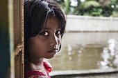 Noakhali, Bangladesh - 02 September 2024: View of a young girl's intense gaze, framed by the weathered doorway and the soft, diffused light reflecting off the water.