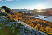 Aerial view of a solitary figure stands atop a rugged, moss-covered peak, overlooking the tranquil waters and distant cityscape of Svolvær, Nordland, Norway.