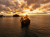 Aerial view of a small island with palm trees silhouetted against the setting sun, casting a golden glow over the tranquil waters, Nuquí, Choco, Colombia.