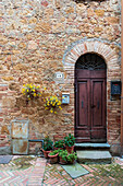 Tuscany, Italy - 22 April 2025: View of a rustic stone wall embracing a weathered wooden door, adorned with vibrant yellow flowers and potted plants.