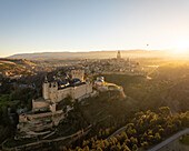 Luftaufnahme des Alcázar de Segovia, wie er erhaben dasteht, während goldenes Sonnenlicht die alten Steine und die umliegende Landschaft eintaucht, Segovia, Kastilien und León, Spanien.