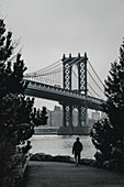 New York, Vereinigte Staaten - 25. Dezember 2023: Blick auf die Manhattan Bridge, während eine einsame Gestalt auf den East River zugeht, eingerahmt von den dunklen Silhouetten der Bäume.