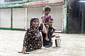 Feni, Bangladesh - 21 August 2024: View of a family wading through floodwaters near closed shops, their determination etched against the backdrop of the submerged landscape.