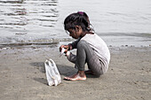 Cox's Bazar, Bangladesh - 01 January 2000: View of a child crouching on the sandy shore, a plastic bag beside her, as she plays by the water's edge.