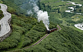 Luftaufnahme eines Oldtimerzuges, der weiße Wolken aufwirbelt, während er sich durch üppig grüne Hügel entlang eines gewundenen Flusses und einer kurvenreichen Straße schlängelt, Furkapass, Schweiz.