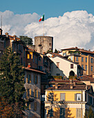 View of sunlit buildings cascade down the hillside, crowned by an ancient stone tower waving the Italian flag against a backdrop of fluffy clouds, Bergamo, Lombardy, Italy.