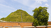 View of a vibrant green mound stands tall behind a weathered brick wall, crowned with a flag under a bright sky, Kraków, Lesser Poland Voivodeship, Poland.