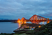 Blick auf die Forth Bridge, eine freitragende Eisenbahnbrücke, die in der Abenddämmerung orange leuchtet und sich im ruhigen blauen Wasser des Firth of Forth, North Queensferry, Schottland, spiegelt.