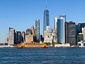 Blick auf eine leuchtend orangefarbene Fähre, die durch das tiefblaue Wasser fährt, vor dem Hintergrund hoch aufragender Wolkenkratzer unter einem klaren Himmel, New York, New York, Vereinigte Staaten.