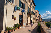 View of sun-drenched, aged buildings line a terracotta street where laundry hangs overhead and a lone woman strolls, Pienza, Tuscany, Italy.