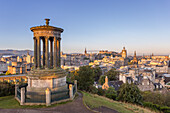 View of the iconic Dugald Stewart Monument stands majestically on Calton Hill, overlooking the sprawling cityscape basking in the warm glow of the setting sun, Edinburgh, Scotland.