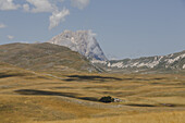 Blick auf goldene Felder, die sich in Richtung des majestätischen Gran Sasso erstrecken, dessen Gipfel in sanfte Wolken gehüllt ist und Schatten wirft, in Isola del Gran Sasso D'italia, Abruzzen, Italien.