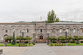 View of aged stone building with arched windows and a small flag fluttering atop, surrounded by verdant trees and benches, Goris, Syunik Province, Armenia.