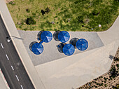 Aerial view of vibrant blue umbrellas casting playful shadows on the geometrically patterned pavement, contrasted by the smooth asphalt road, Nahariyya, North District, Israel.