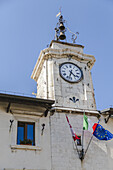 View of an ornate clock tower under a clear blue sky, adorned with flags, a bell, and intricate stone carvings, standing tall in Pescocostanzo, Abruzzo, Italy.