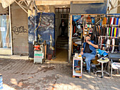 Jerusalem, Israel - 28. Oktober 2019: Blick auf einen Mann, der vor einem Geschäft sitzt, umgeben von farbenfrohen Lederwaren und Werkzeugen, die einen lebhaften Kontrast zu den verwitterten Wänden und dem ruhigen Eingang hinter ihm bilden.