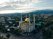 Luftaufnahme der Baiken-Moschee mit ihrer goldenen Kuppel inmitten der Gebäude der Stadt und vor dem Hintergrund der schneebedeckten Berge, Almaty, Region Almaty, Kasachstan.