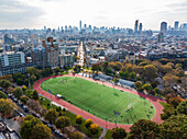 Aerial view of a vibrant green sports field encircled by a red track, nestled among autumnal trees and urban buildings stretching to the distant skyline, Brooklyn, New York, United States.