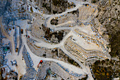 Luftaufnahme einer kahlen, terrassenförmig angelegten Landschaft, die von Marmorbrüchen geformt wurde und geometrische Muster und verschlungene Straßen erkennen lässt, ein Zeugnis menschlicher Industrie, Carrara, Toskana, Italien.