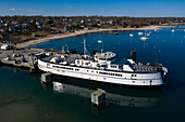 Aerial view of a white ferry docked at a pier, the calm water reflecting the blue sky and the distant shore lined with trees, Martha's Vineyard, Massachusetts, United States.