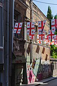 View of Georgian flags flutter proudly above a weathered building with graffiti-covered walls, casting shadows on the street, Tbilisi, Tbilisi, Georgia.