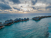 Malediven, Malediven - 28. Mai 2019: Luftaufnahme der Überwasser-Bungalows eines luxuriösen Resorts, die durch Holzstege miteinander verbunden sind, die vor dem türkisfarbenen Wasser und dem Himmel in den sanften Farben der Morgendämmerung schimmern.