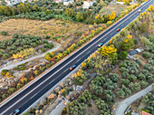 Aerial view of a long road cutting through the landscape, lined with vibrant greenery, houses and multiple cars, Chania, Chania, Greece.