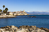 View of the old town with its historic walls, sandy beaches, and the shimmering turquoise sea against the backdrop of snow-capped mountains, Antibes, Provence-Alpes-Côte d'Azur, France.