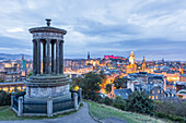 View of the monument atop Calton Hill overlooking the city's skyline with the castle illuminated in pink against the twilight sky, Edinburgh, Scotland.