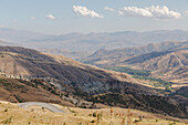 View of sun-drenched hillsides cascading into a valley, a winding road cutting through the golden terrain under a sky dotted with clouds, Vardenyats Pass, Armenia.