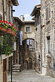View of narrow cobblestone streets lined with weathered stone buildings, bright red flowers cascading from balconies, and laundry hanging overhead, Scanno, Abruzzo, Italy.