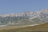Blick auf das majestätische, schneebedeckte Gran-Sasso-Gebirge, das sich scharf gegen den klaren blauen Himmel abhebt und Schatten auf die Grasflächen darunter wirft, Isola del Gran Sasso D'italia, Abruzzen, Italien.