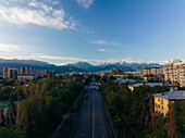 Aerial view of a road stretching towards the snow-capped mountains beyond the city's buildings and green canopy, Almaty, Almaty Region, Kazakhstan.