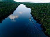Aerial view of a Churún river reflecting the sky, flanked by dense green jungle, with a boat leaving a white wake through the dark water, Canaima National Park, Bolívar, Venezuela.