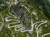 Aerial view of a winding road snaking through rugged, rocky terrain, a contrast of grey stone and green grass, Gotthard Pass, Ticino, Switzerland.
