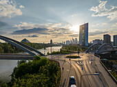 Aerial view of the sun blazing through the glass facade of the European Central Bank tower, reflected in the Main River, with bridges arching over lush greenery, Frankfurt am Main, Hessen, Germany.