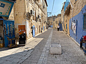 Jerusalem, Israel - 30 October 2019: View of a narrow street lined with aged buildings, the stone pavement reflecting warm sunlight between the blue accents of doors and decorations.