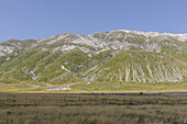 View of rolling green hills cascade towards a golden field under a serene blue sky, the mountains capped with white, Isola del Gran Sasso D'italia, Abruzzo, Italy.