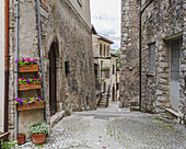 View of aged stone buildings stand guard over a cobblestone street, flowers spilling from window boxes, in the heart of Scanno, Abruzzo, Italy.