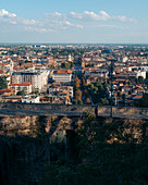 View of sun-kissed rooftops sprawling towards the horizon, framed by ancient stone walls under a vast blue sky, Bergamo, Lombardy, Italy.