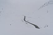 Aerial view of a winding road cutting through a stark, snow-covered landscape, the pass shrouded in a soft, ethereal mist, Julier Pass, Grisons, Switzerland.