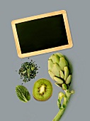 Grünes Stillleben (Artischocke, Kiwi, Kräuter) mit schwarzer Schiefertafel