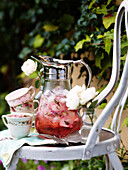 Mit Rosenblättern aromatisierter Punsch im Glaskrug auf Stuhl im sommerlichen Garten