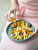 Woman pouring orange sauce over a dish of roasted root vegetable, feta and squash seed salad