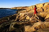 Woman jumping over rocks on coast, Bohuslan region, Vastra Gotaland, Sweden