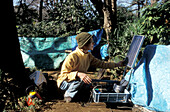 Homeless, living boxes in Tokyo, Japan, Homeless community in Ueno Park woman checking her solar panel for electricity of her tiny TV in a basic tent Obdachlose, notdürftige Schutzbauten, Pappkarton-Architektur, Plastikplanen, Slum, Obdachlosigkeit, Randg