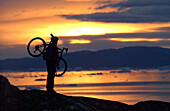 Mountainbiker trägt sein Fahrrad, betrachtet den Aussicht beim Sonnenuntergang, Ilulissat, Greenland