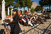 People sitting in deck chairs at Museumshafen, People sitting in deck chairs of an open-air cafe at sandy beach of Museumshafen oevelgoenne while waitress serving coffee, Hamburg, Germany