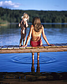 Mother and daughter sitting on jetty, dangling feet in water, rear view